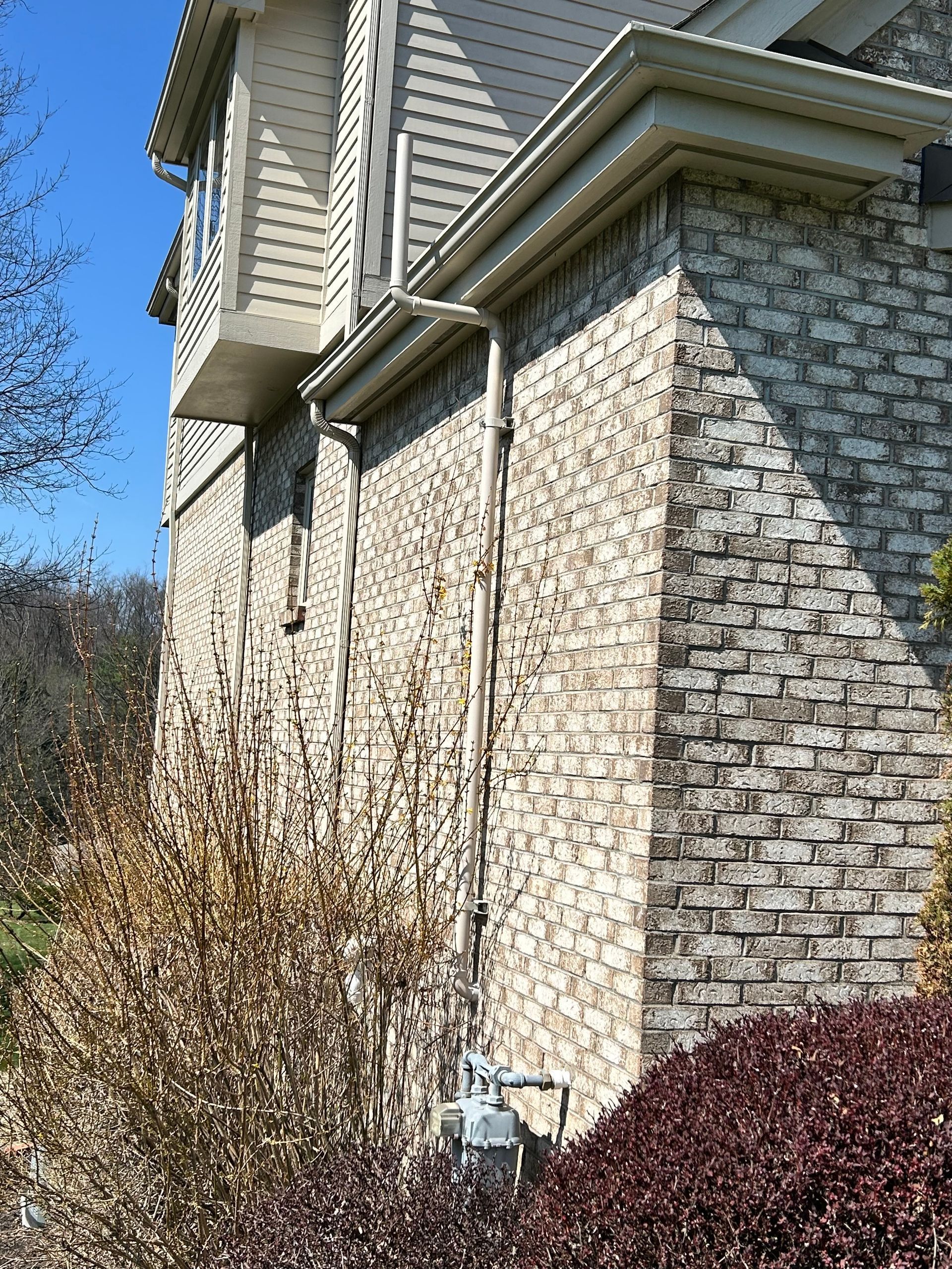 A beige brick wall of a house with a vertical metal gas pipe running up the side to a meter near the ground.