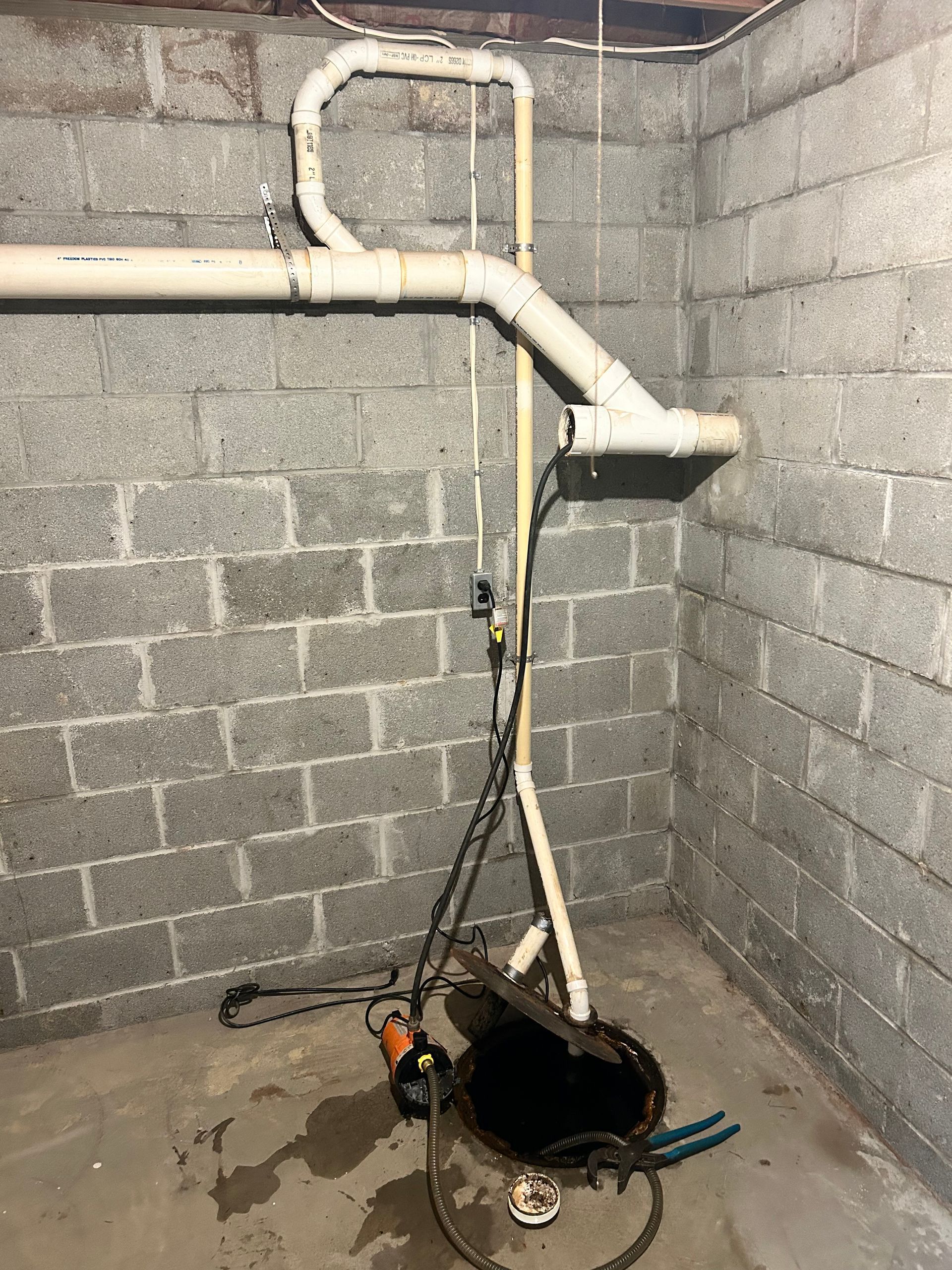 A sump pump system in a basement with PVC piping connected to an open, black drainage pit against a cinder block wall.