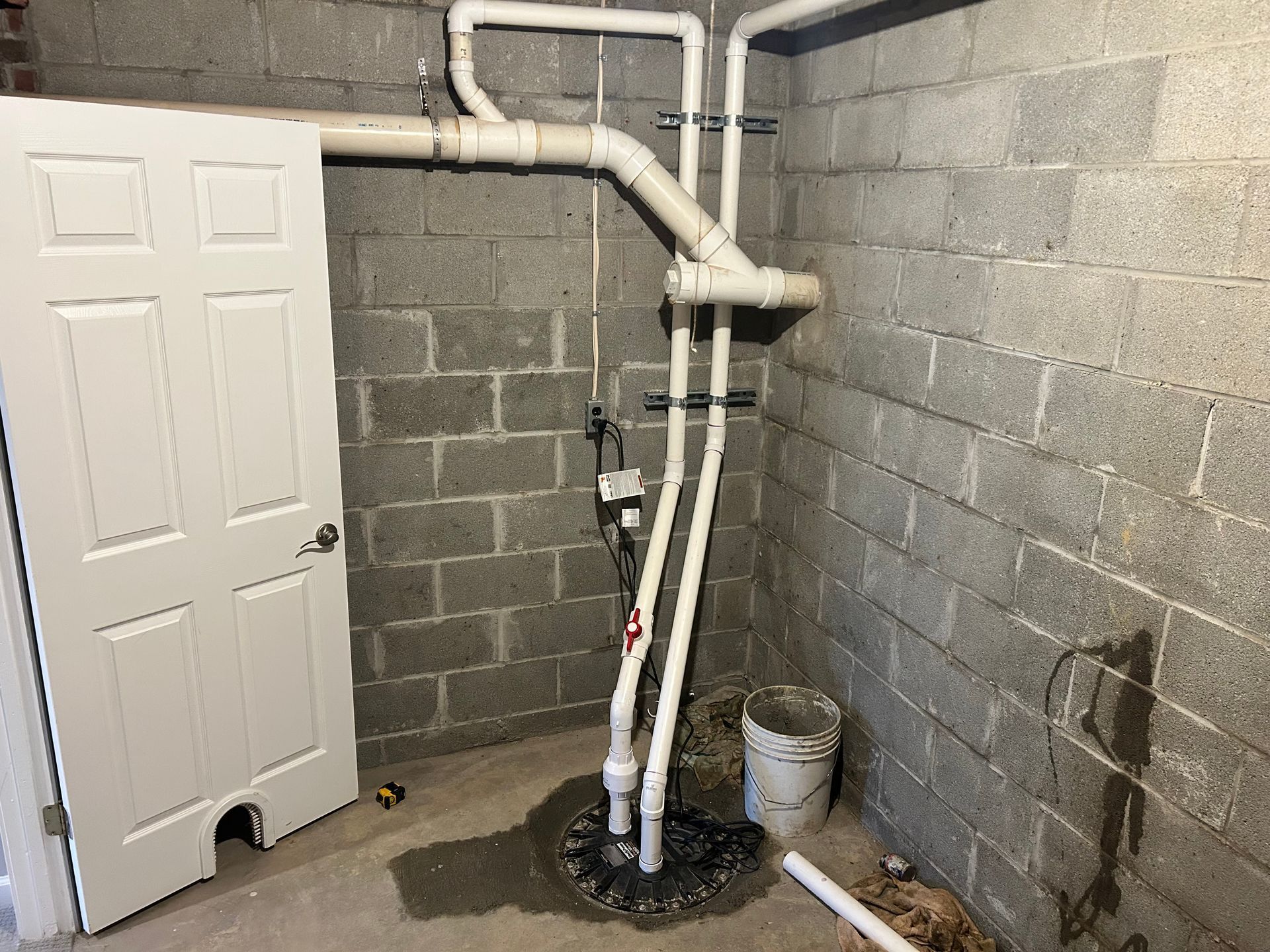 A sump pump with white PVC piping installed in a grey concrete block corner next to a white door.