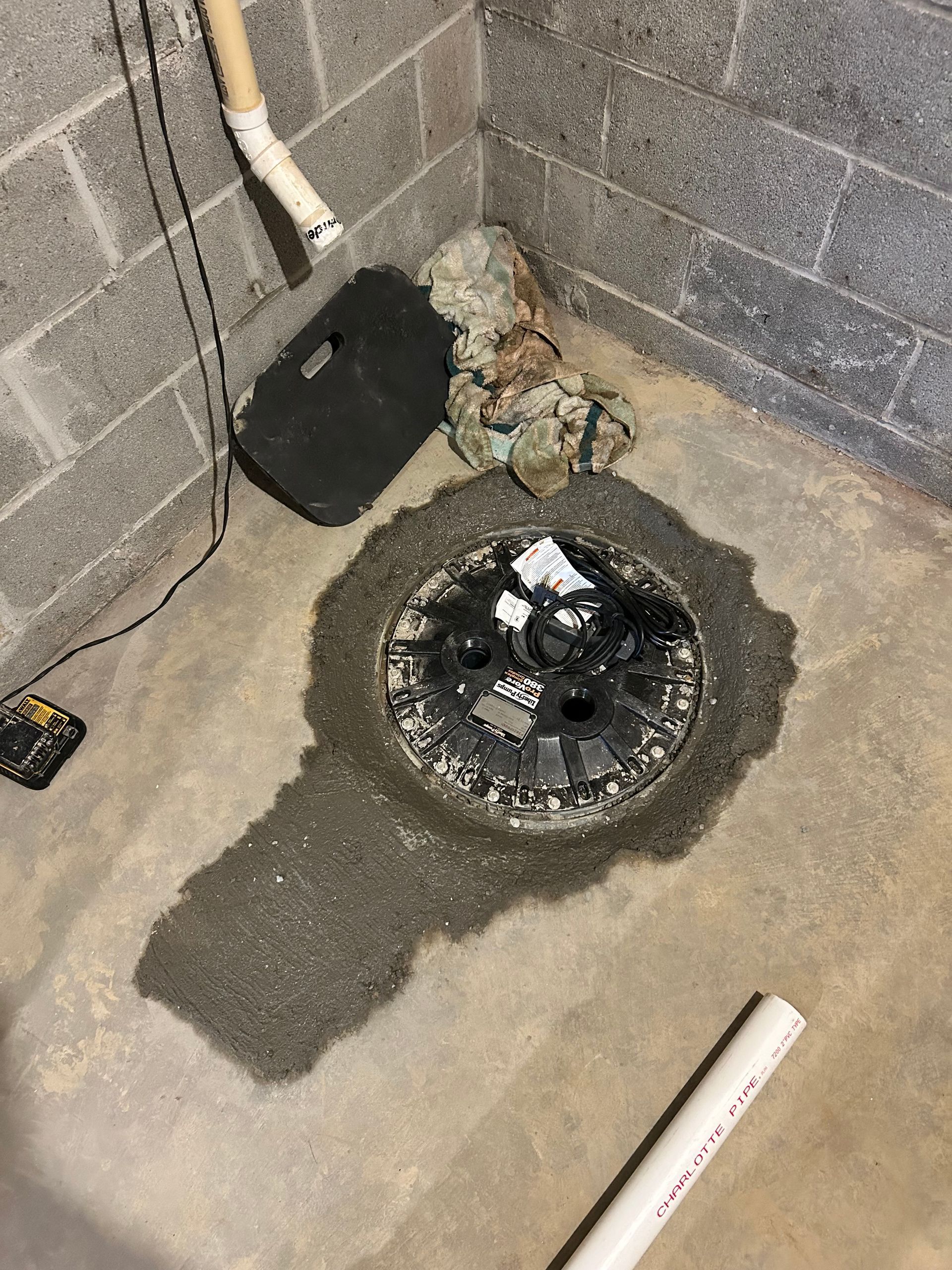 A circular sump pump lid set into a concrete basement floor, surrounded by recently applied mortar or concrete patch.