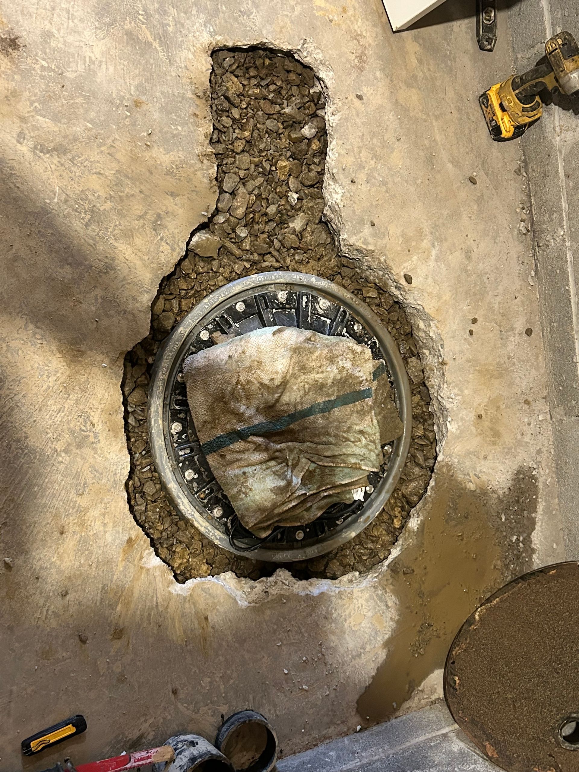 A circular manhole access point set into concrete, surrounded by gravel and repair tools.