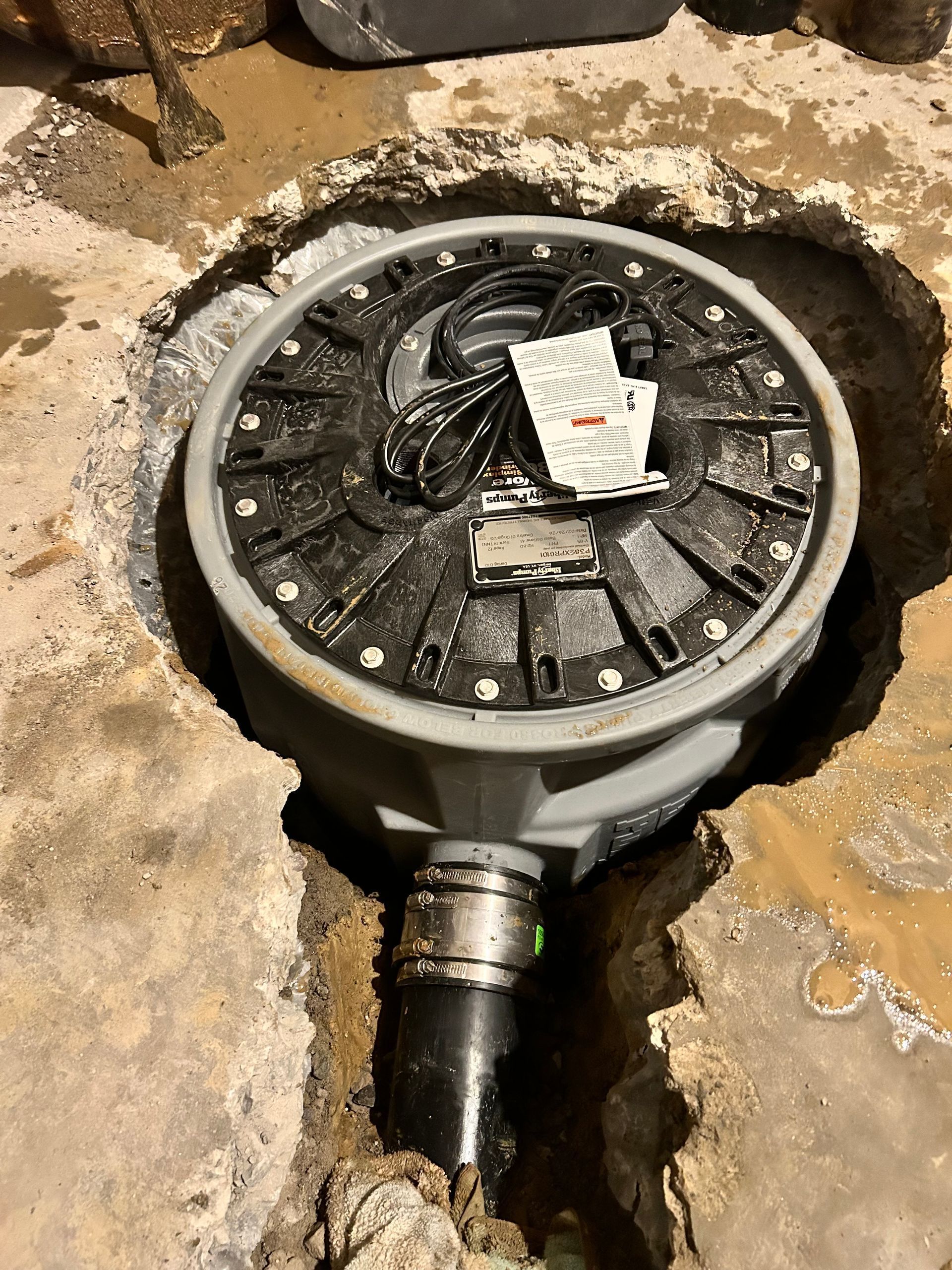 A sewage ejector pump installed in a hole in the ground, showing the black circular lid and connected black pipe.