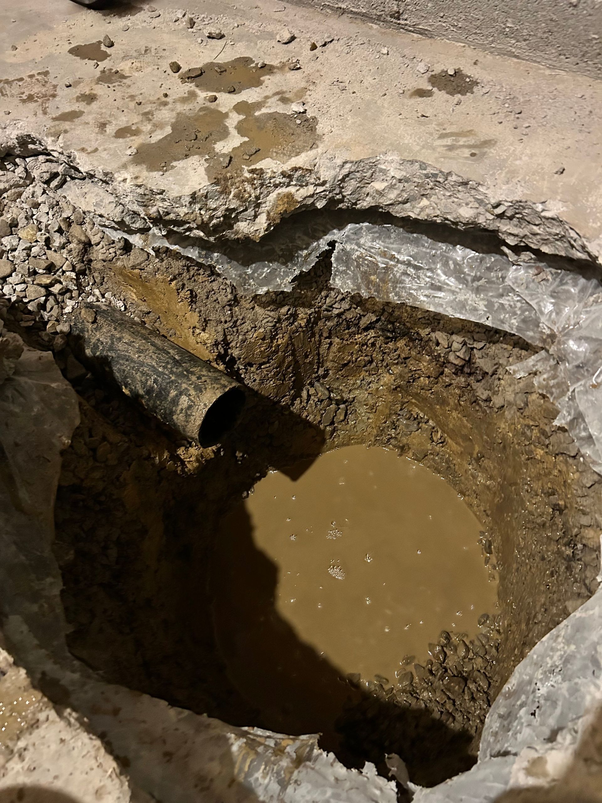 An open hole in a paved area, showing a pipe protruding into the side of a dirt pit filled with muddy water.
