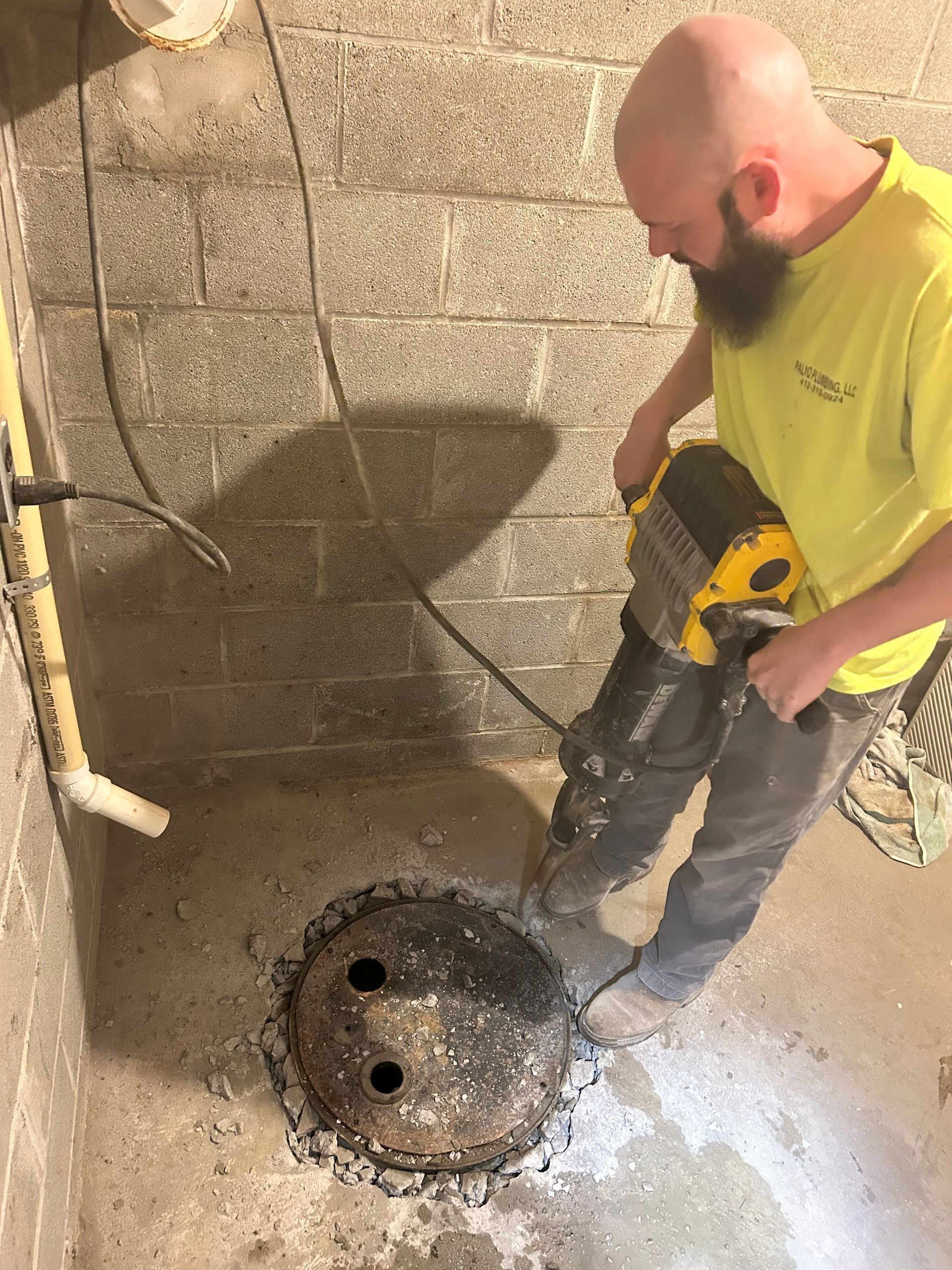 A person in a yellow shirt uses a jackhammer to break up concrete in a basement floor around a drain cover.