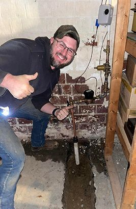 A smiling person in work clothes kneels in a basement, giving a thumbs-up next to new residential water plumbing.