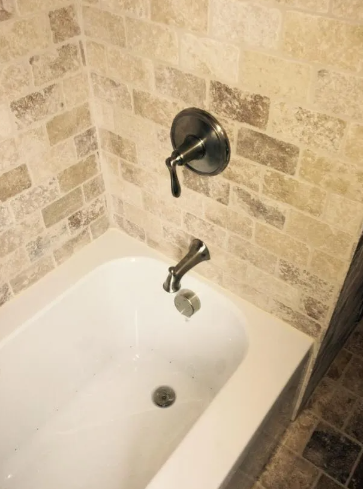 A bathtub with a bronze-toned faucet, shower handle, and drain set against a neutral, brick-patterned tiled wall.