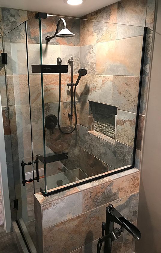 A walk-in shower with rustic, multi-colored stone wall tiles, black fixtures, a glass door, and a built-in recessed shelf.