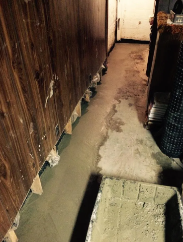 Fresh concrete floor patch along a wooden-paneled basement wall, with a cement mixing tub in the foreground.