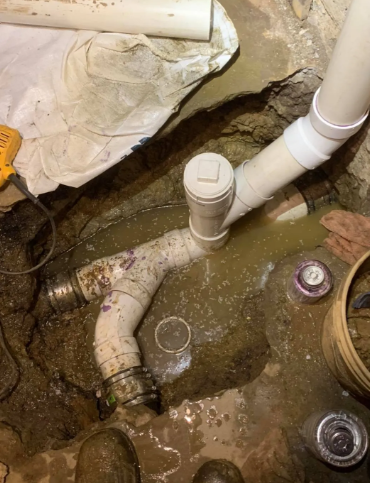 White PVC plumbing pipes joined with a T-fitting in a muddy, excavated trench in the ground.