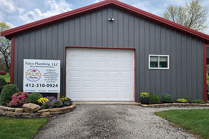 Gray metal garage building with a red roof, white door, a sign for Palvo Plumbing, and a landscaped flower bed in front.