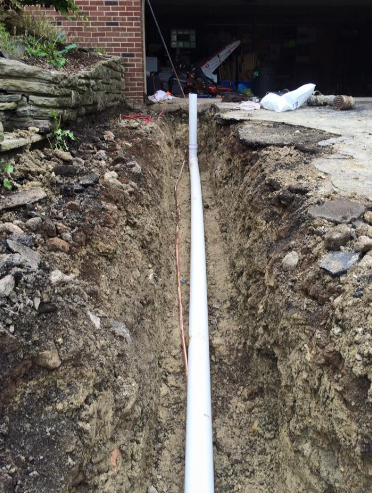 A white PVC pipe runs through a freshly dug trench next to a concrete driveway and a brick wall.