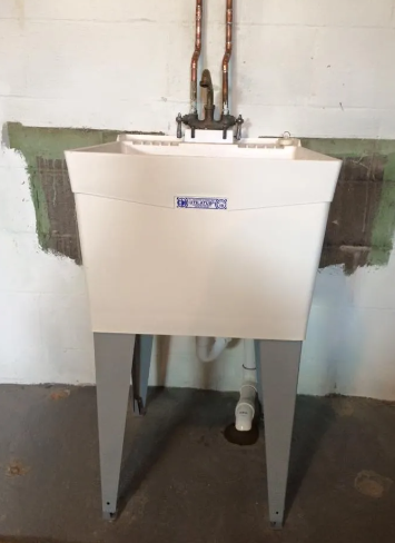 A white utility sink with a faucet stands on metal legs against a white block wall with exposed copper pipes.