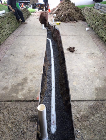 A trench cut through a concrete walkway shows a white pipe being installed over a gravel base for drainage.