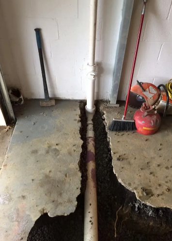 A vertical PVC sewer pipe runs through a trench cut into a concrete garage floor, surrounded by construction tools.