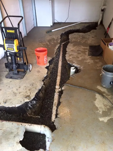 Concrete floor opened with a trench exposing buried white plumbing pipes, with a tool and buckets nearby in a garage.