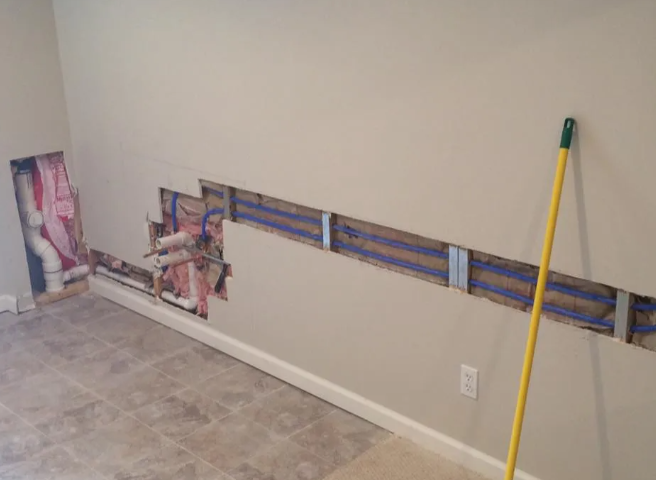 A wall with a rectangular drywall section removed, revealing blue plumbing pipes, wooden studs, and a vertical drain pipe.