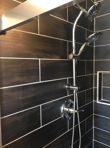 Chrome shower fixture with a fixed head and a handheld sprayer against dark, horizontal rectangular tiled walls.