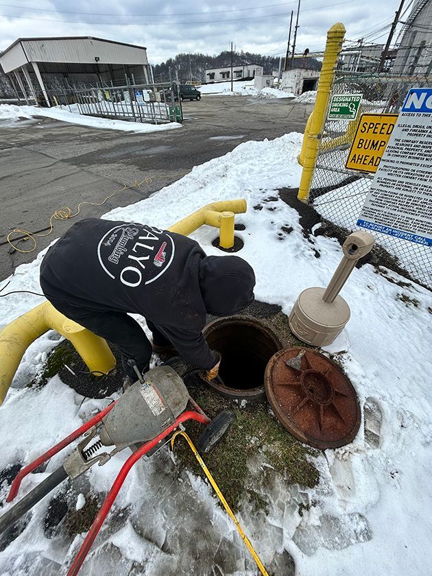A worker in a black hoodie inspects an open sewer manhole in a snowy outdoor industrial area.