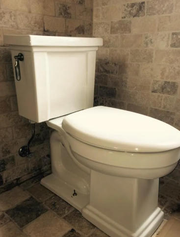 A white toilet with an elongated seat in a bathroom featuring rustic, multi-toned stone tile walls and flooring.