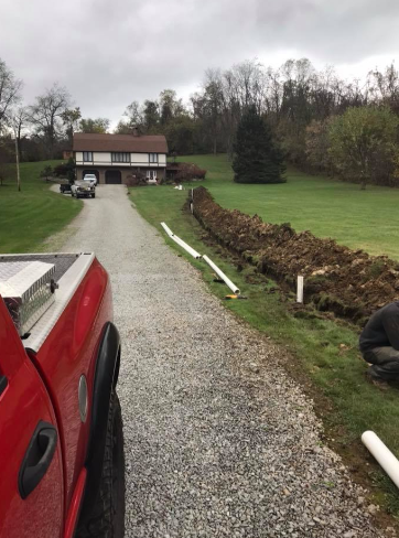 A red truck is parked on a gravel driveway next to a long, dug-up trench in a grassy yard near a house.