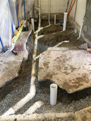 White PVC plumbing pipes installed in a trench dug through a concrete basement floor, surrounded by gravel.