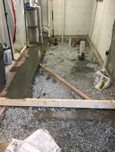 A basement under construction featuring a concrete floor patch, gravel ground, exposed piping, and wooden framing boards.