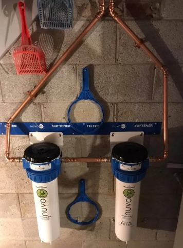 Two Nuvo water filter housings mounted on a cinder block wall, connected by copper piping and blue wall brackets.