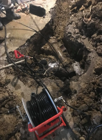 A camera inspection reel sits on dirt next to an open trench revealing exposed underground pipes.