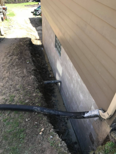 A trench dug along the side of a tan house foundation, with black drainage pipes extending from the wall and ground.