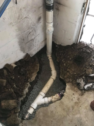 Newly installed white PVC plumbing pipes in a dirt-filled trench inside a basement corner.