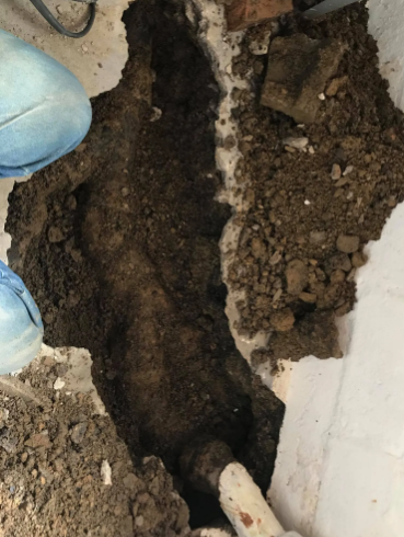 A trench dug through a concrete floor exposing dark soil and a white PVC pipe.