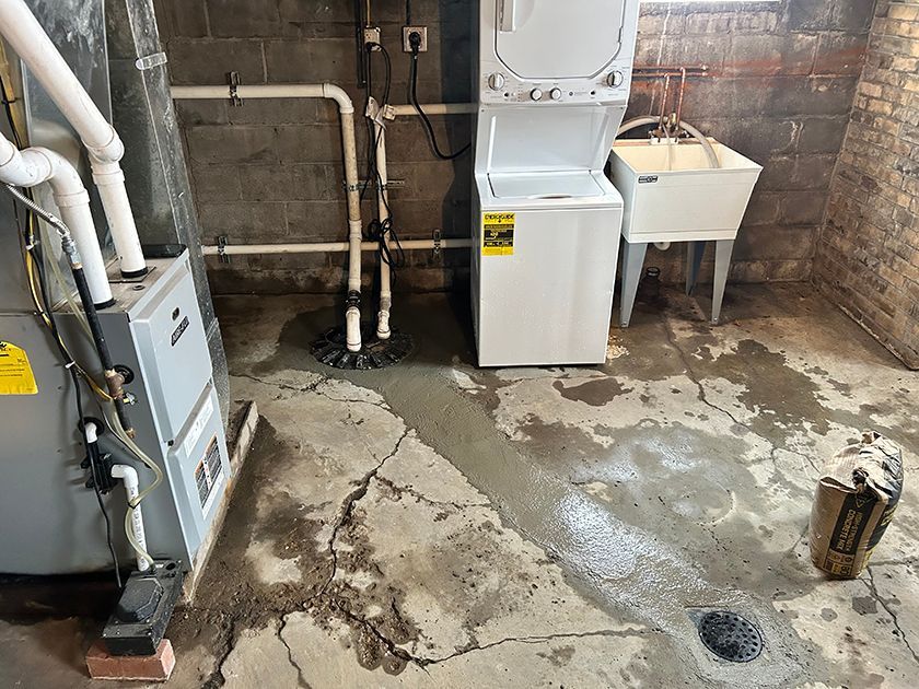 A basement floor with fresh concrete patches connecting a sump pump area to a drain, near a laundry unit and utility sink.