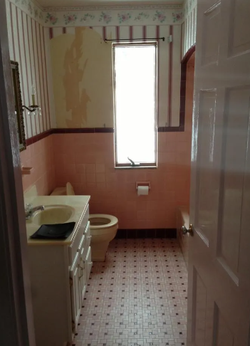 A bathroom features pink tiled walls, striped wallpaper with a peeling section, a vanity, toilet, and patterned tile floor.