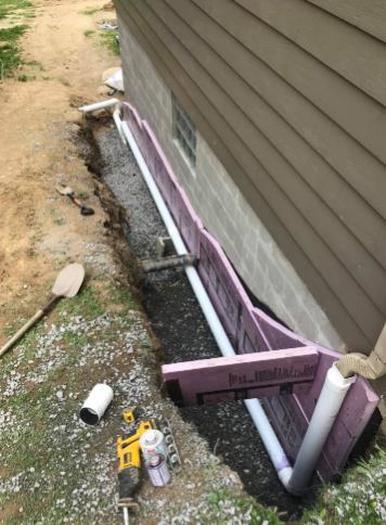 A trench along a house foundation containing a drainage pipe, gravel, and pink foam insulation boards.