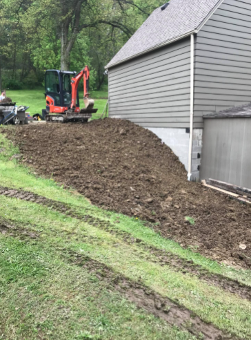 An orange mini-excavator parked on a freshly graded dirt mound next to the gray siding and white foundation of a house.