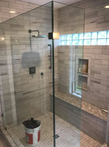 A glass-enclosed shower with light-colored brick-style wall tiles, a built-in stone bench, and a bucket on the floor.