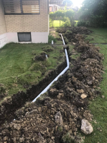 A grey PVC drain pipe runs through an open trench in a residential yard beside a brick house.