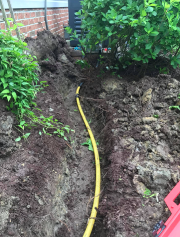 A bright yellow gas line runs through a narrow trench in the dirt, alongside a brick wall and green garden bushes.