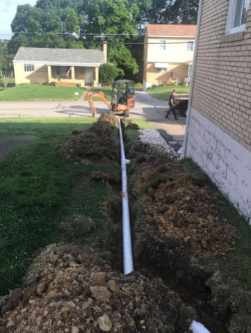 A white PVC pipe lies in an excavated trench alongside a brick house, with a small excavator visible in the background.