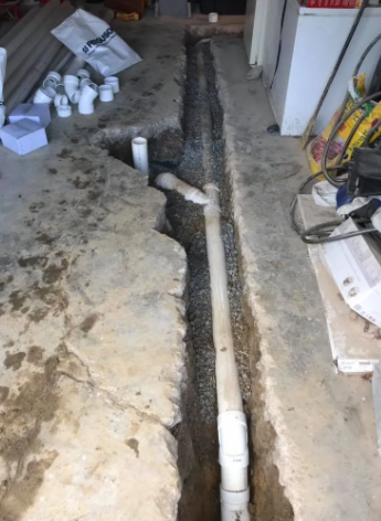 A white PVC pipe system installed in a narrow trench cut through a concrete floor, surrounded by plumbing supplies.