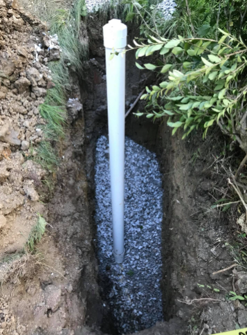 A vertical PVC pipe stands inside a narrow, gravel-filled trench in the ground beside some bushes.