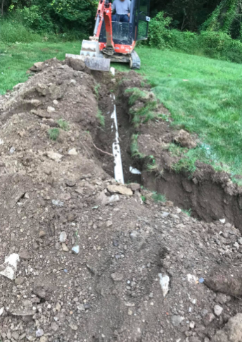 An orange excavator digs a trench in a grassy yard, revealing a white pipe laid in the ground.