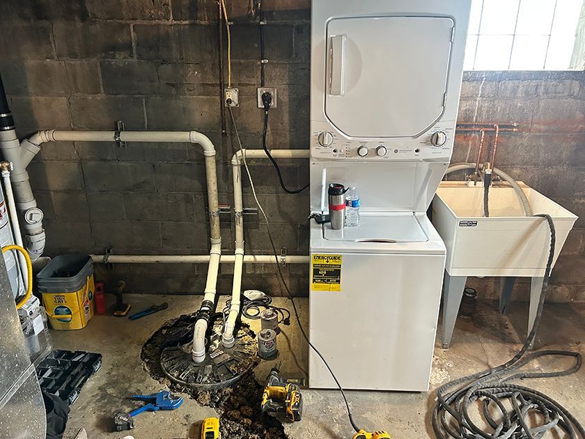 A white stacked washer-dryer unit stands next to a utility sink and a sump pump in a basement with exposed block walls.