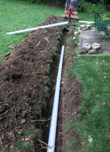 A white drainage pipe laid in a narrow dirt trench in a backyard, with a small excavator visible in the background.