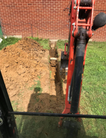 View from an excavator cab showing an orange mechanical arm digging a long trench in a grassy yard beside a brick wall.