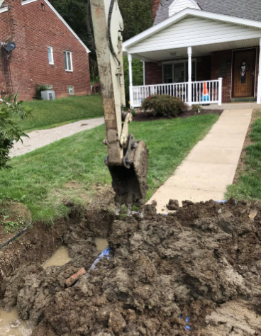 An excavator bucket suspended over an excavated dirt trench in front of a house.