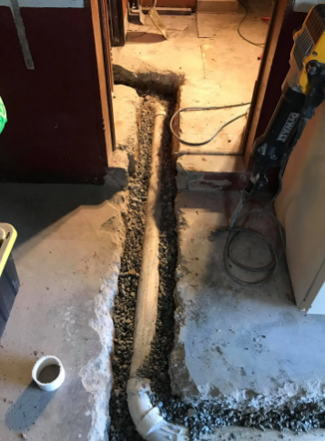 A white PVC pipe lies in a trench cut into a concrete floor, surrounded by gravel, with a power tool nearby.