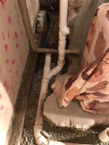 White PVC plumbing pipes installed in a narrow dirt trench alongside a concrete foundation and a covered appliance.