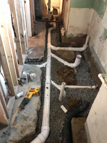 A trench cut into a concrete floor reveals a new white PVC plumbing system and gravel bedding under construction.