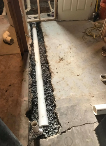 A white PVC drain pipe sits in a gravel-filled trench cut into a concrete floor during a construction project.